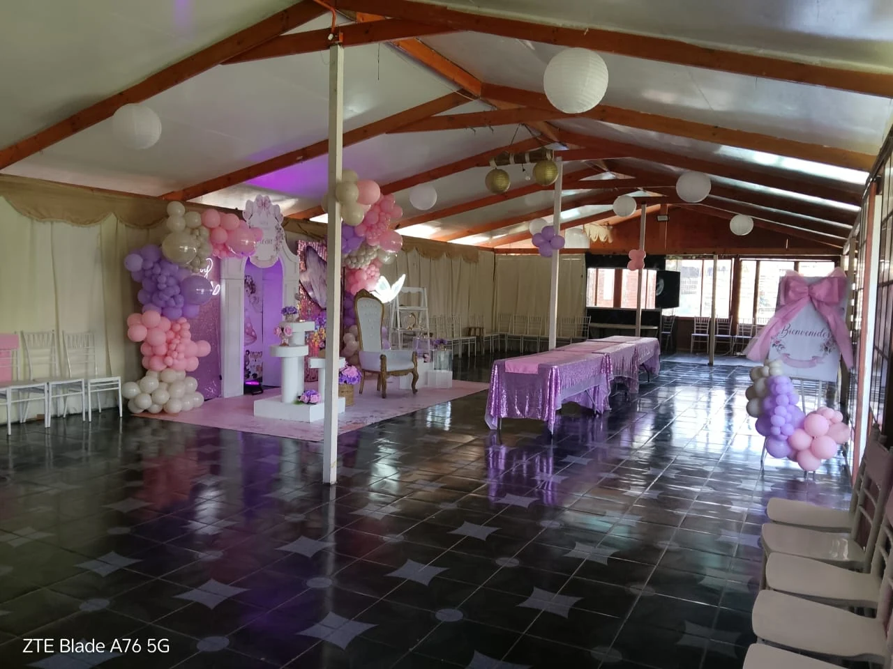 Vista completa del salón preparado para un baby shower con decoración rosa y lila, mesas alargadas y sillas blancas.