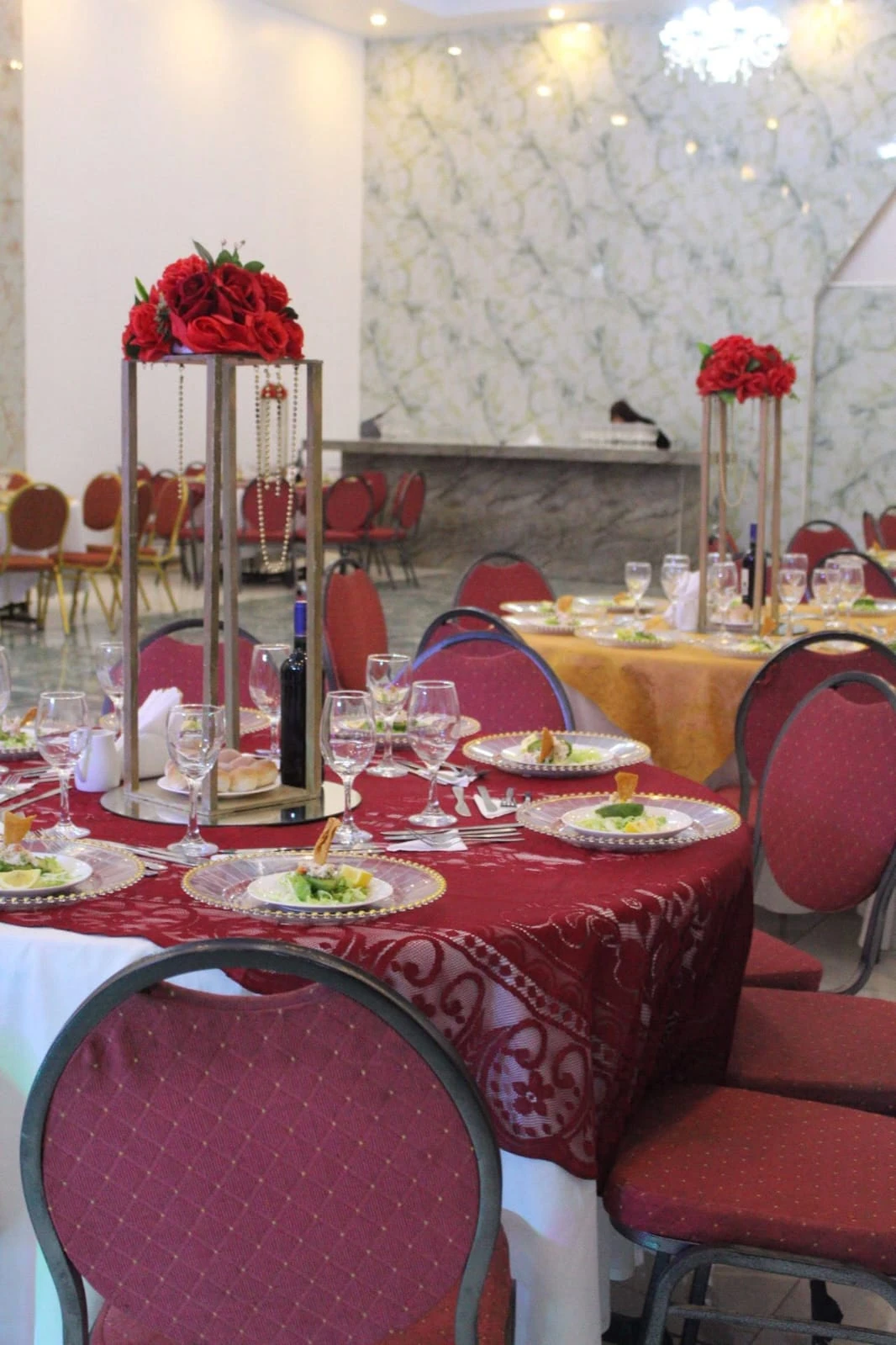 Mesas de cena corporativa con mantelería burdeos y dorada, platos servidos, copas y centros florales rojos altos.