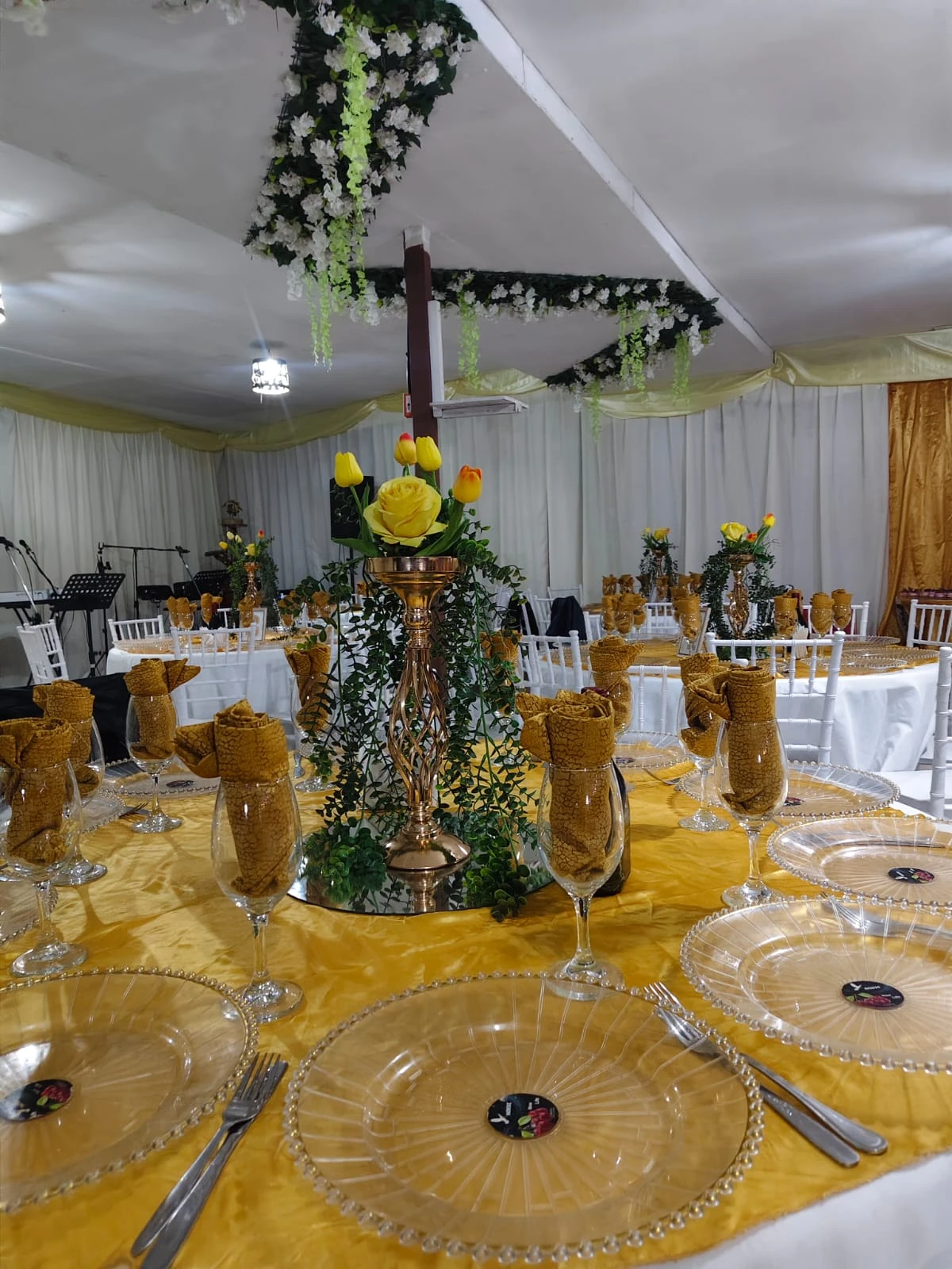 Mesa de Manantial con mantelería dorada, copas, platos transparentes y un centro floral alto con rosas y tulipanes amarillos.