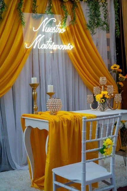 Mesa de matrimonio con telas doradas y blancas, girasoles, velas y un letrero luminoso que dice Nuestro Matrimonio.
