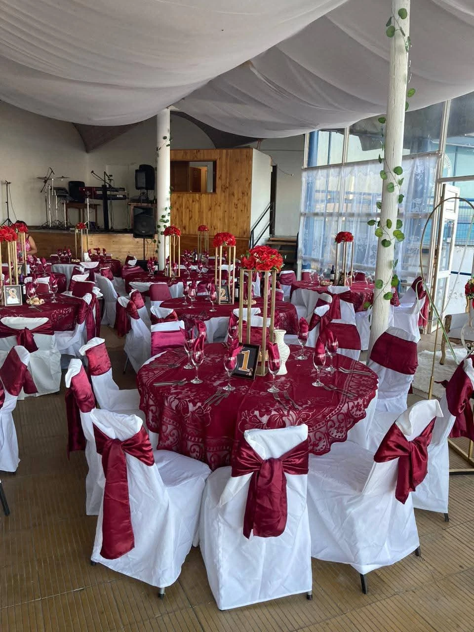 Salón preparado para una cena corporativa con mesas redondas en burdeos, sillas blancas con lazos y centros florales rojos.