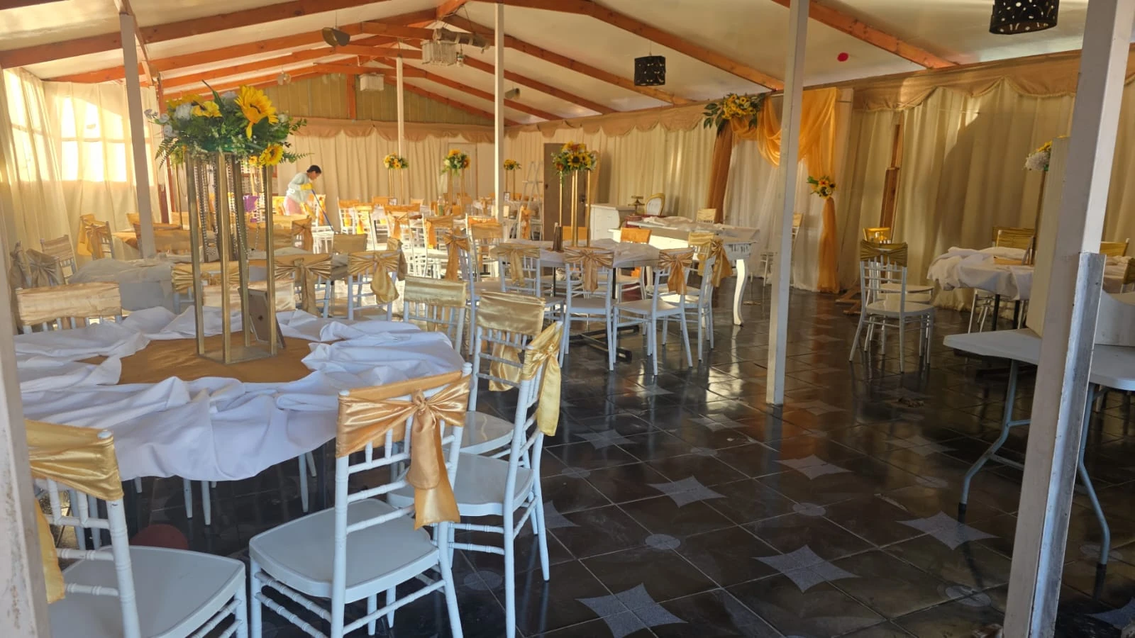 Salón de Manantial preparado con mesas claras, sillas doradas y centros florales bajo vigas de madera.