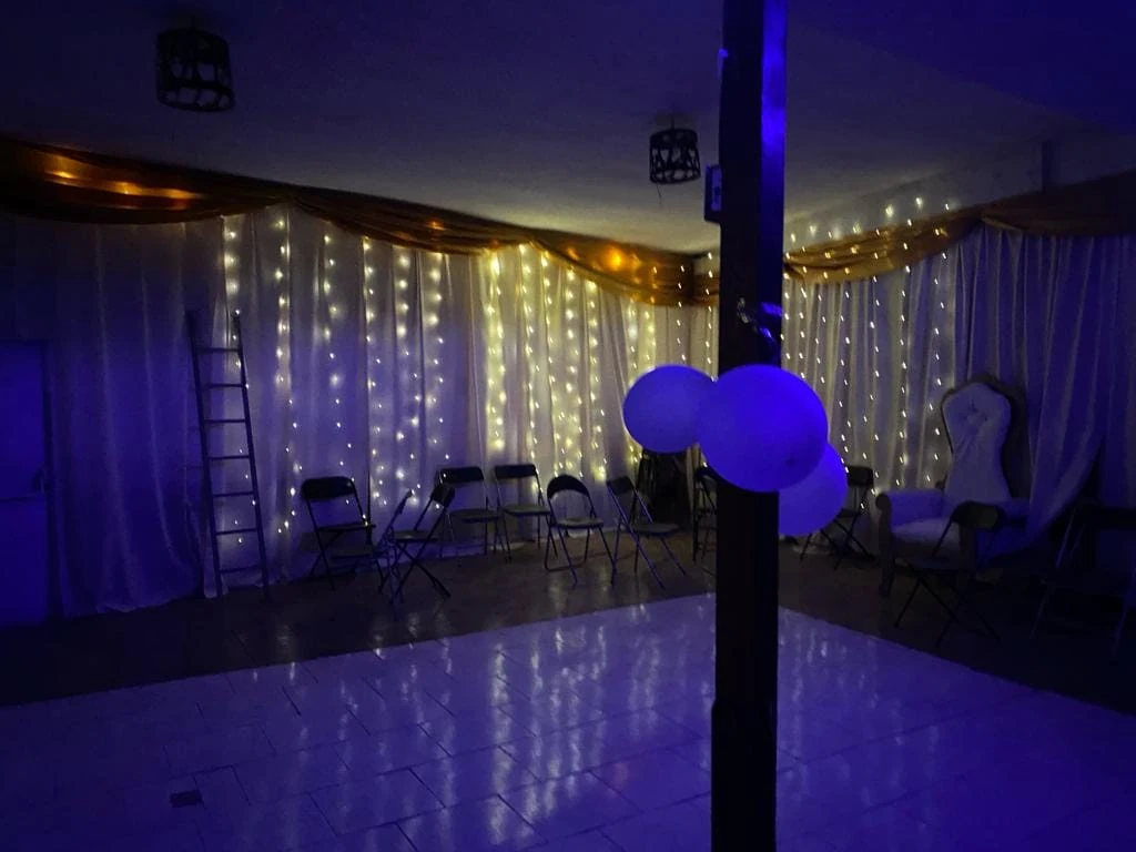 Interior de un salón de Manantial con cortinas claras, guirnaldas luminosas, globos azules y luz violeta.